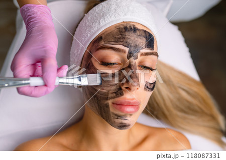 cosmetologist applying black mask on pretty woman face wearing black gloves, gorgeous woman in spa having facial procedures 118087331