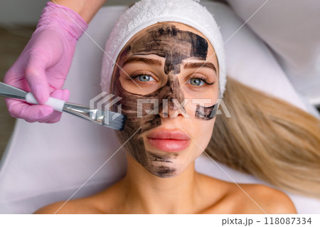 cosmetologist applying black mask on pretty woman face wearing black gloves, gorgeous woman in spa having facial procedures 118087334
