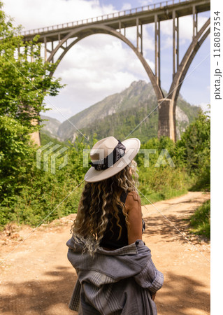A girl in a hat stands against the backdrop of a beautiful arched bridge over the Tara River, a popular tourist attraction in Montenegro. 118087354