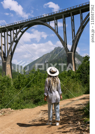 A girl in a hat stands against the backdrop of a beautiful arched bridge over the Tara River, a popular tourist attraction in Montenegro. A girl in a hat stands against the backdrop of a beautiful arched bridge over the Tara River, a popular tourist attraction in Montenegro. 118087362