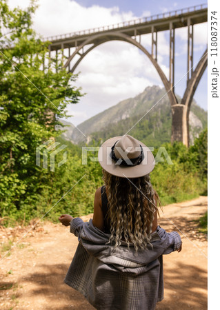 A girl in a hat stands against the backdrop of a beautiful arched bridge over the Tara River, a popular tourist attraction in Montenegro. A girl in a hat stands against the backdrop of a beautiful arched bridge over the Tara River, a popular tourist attraction in Montenegro. 118087374