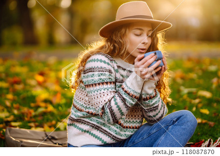 Curly woman with thermos in clearing among fallen leaves in autumn park, enjoying hot drink. 118087870