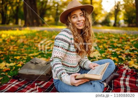 Beautiful curly woman in hat on mat with book in autumn park. Relaxation, solitude with nature. Beautiful curly woman in hat on mat with book in autumn park. Relaxation, solitude with nature. 118088020