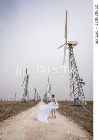 A woman in a white dress is walking down a dirt road in front of a row of wind turbines. 118088062