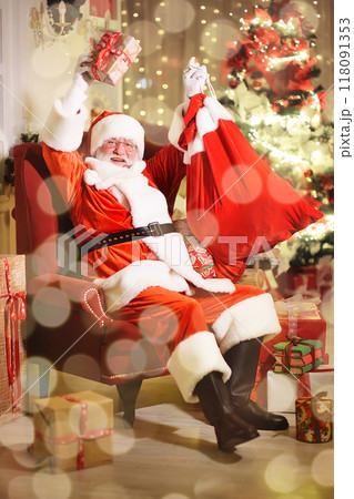 Santa Claus is smiling sitting on a red armchair and holding a bag of gifts against the background of a Christmas tree and Christmas lights. 118091353