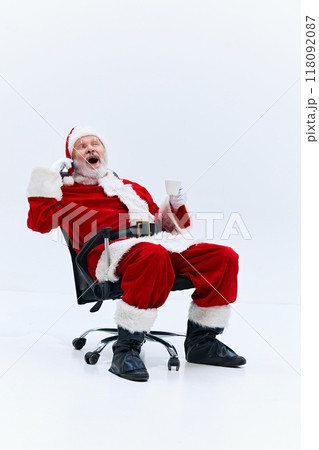 Santa Claus sitting on rolling office chair, holding cup in one hand, and talking one phone with excited expression against white background. Santa Claus sitting on rolling office chair, holding cup in one hand, and talking one phone with excited expression against white background. 118092087