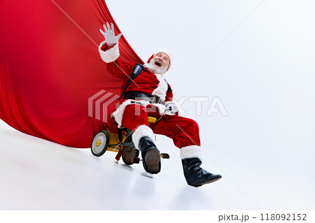 Santa Claus on small tricycle, holding red bag of gifts behind him, with joyful expression, against white studio background. 118092152
