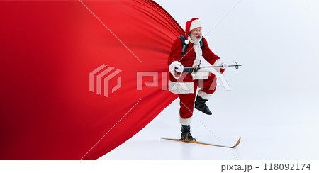 anta Claus balancing on one ski with a large red cape flowing behind, holding ski pole, mid-motion, against white studio background. 118092174