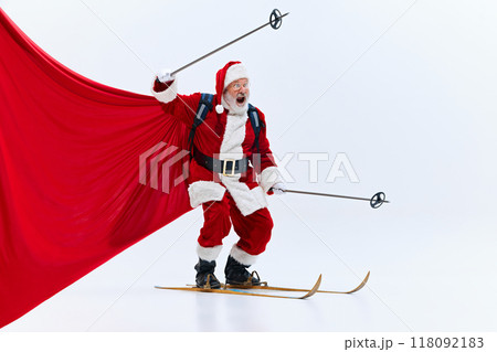 Santa Claus, shouting with enthusiasm, gripping steering wheel, skiing with red cape behind against white studio background. Santa Claus, shouting with enthusiasm, gripping steering wheel, skiing with red cape behind against white studio background. 118092183