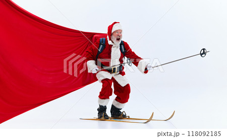 Santa Claus, smiling excitedly, holding steering wheel while skiing, red fabric trailing behind, against white studio background. 118092185