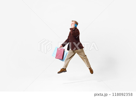 Stylish young man wearing beige pants and brown jacket, in motion with colorful shopping bags, talking on phone. Excitement 118092358
