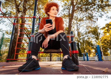 Bottom view of mid adult Caucasian tired woman sitting at sports ground and using cell phone. Concept of motivation and relax after training Bottom view of mid adult Caucasian tired woman sitting at sports ground and using cell phone. Concept of motivation and relax after training 118092775