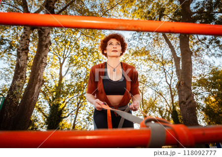 Bottom view through horizontal bar of mid adult Caucasian woman training with sports elastic band. Tree crowns in background. Concept of sport 118092777