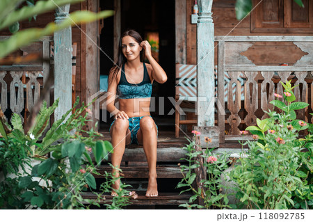 A beautiful tanned woman in summer clothes poses sitting on the wooden steps of a house, surrounded by tropical plants. Summer vacation and travel concept 118092785