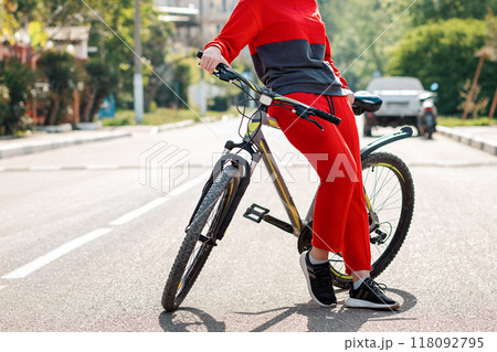 Sports lifestyle. A woman in red sportswear, posing leaning on a Bicycle. In the background, an empty road and street. Eco-friendly transport concept. Close up of legs Sports lifestyle. A woman in red sportswear, posing leaning on a Bicycle. In the background, an empty road and street. Eco-friendly transport concept. Close up of legs 118092795