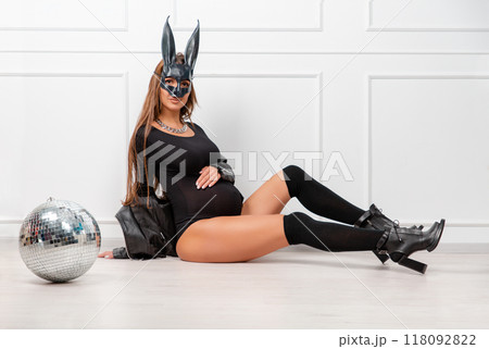 A pregnant woman in a rabbit mask, with long dark hair, in a black bodysuit poses with a mirror ball, holding her stomach with her hands. Studio fashion photography 118092822