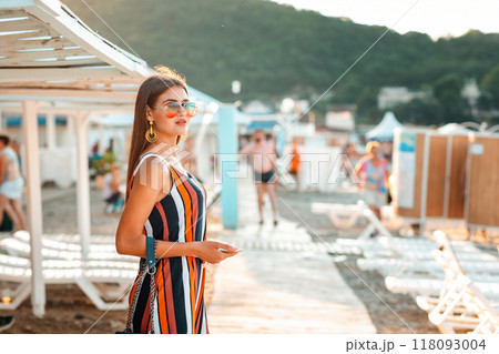 A beautiful woman in a dress and sunglasses stands on the beach. Sun beds and umbrellas are in the background. The concept of summer luxury holidays A beautiful woman in a dress and sunglasses stands on the beach. Sun beds and umbrellas are in the background. The concept of summer luxury holidays 118093004