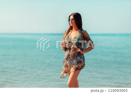 A beautiful woman in a pareo and sunglasses poses against the sea and sky. Concept of beach holidays and summer vacations 118093005