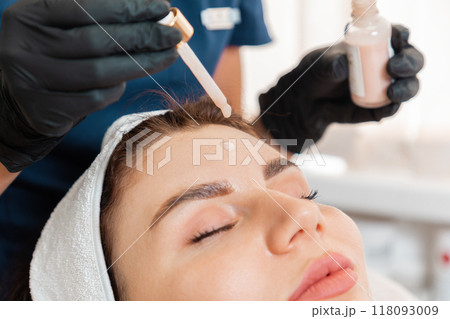 A cosmetologist drips a cosmetic serum on a woman's forehead during the beauty procedure. Close-up portrait of the client. Professional skin care in salon 118093009
