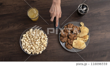 スナックやお菓子のあるテーブル上でリモコンを操作する人。テレビをつける、動画配信を見る、録画を見る 118096559