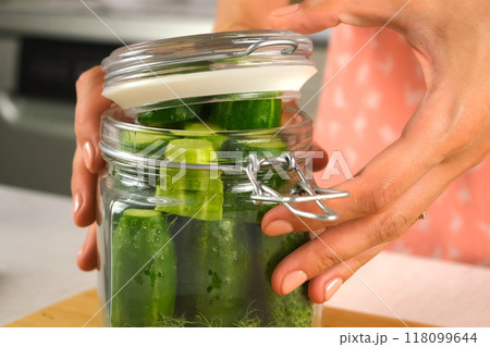 Woman puts cucumbers in glass jar to cook pickles closing lid, hands close-up. 118099644