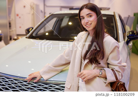 Portrait of successful smiling woman in fashionable beige suit looking at camera and posing next to newly bought car at dealership center, copy space 118100209