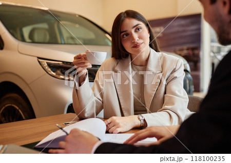 Successful young woman drinking coffee at table while signing purchase contract buying new car at dealership center 118100235