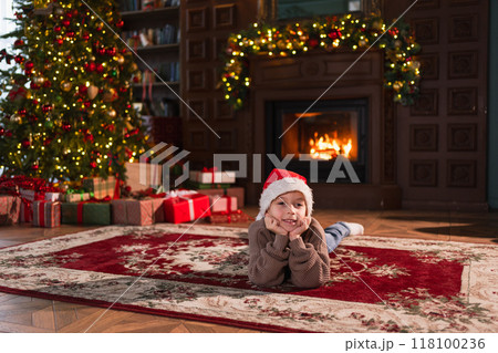 Merry Christmas. Little boy smiling near Christmas tree in classical dark interior. Young happy kid wearing Santa Hat in living room with fireplace Christmas tree gift boxes. Christmas eve at home Merry Christmas. Little boy smiling near Christmas tree in classical dark interior. Young happy kid wearing Santa Hat in living room with fireplace Christmas tree gift boxes. Christmas eve at home 118100236