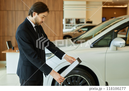 Side view of Central Asian male auto dealer in black suit thoroughly reading information on car dealership stand displaying vehicle spec sheets at dealership center, copy space 118100277