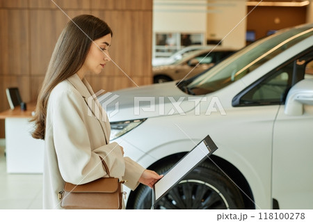 Side view of young woman reading information on car dealership stand displaying vehicle spec sheets at dealership center, copy space 118100278