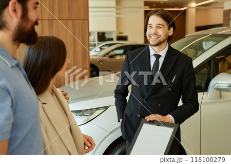 Medium shot of Central Asian male auto dealer talking to couple planning to buy new car. Smiling motor trader assisting clients at dealership center 118100279