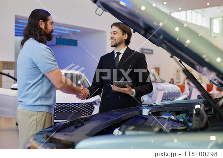 Medium shot smiling Central Asian auto dealer shaking hands with adult man buying new car at dealership center 118100298