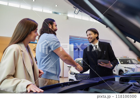 Medium shot smiling Central Asian auto dealer shaking hands with adult man while selling new car to happy couple at dealership center Medium shot smiling Central Asian auto dealer shaking hands with adult man while selling new car to happy couple at dealership center 118100300