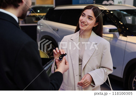 Medium shot of smiling young woman in elegant beige suit talking to auto dealer taking key of new car, while buying vehicle at car dealership center 118100305