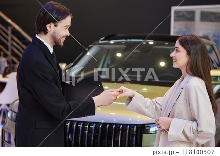 Side view of smiling male auto dealer greeting young female customer planning to buy new vehicle at car dealership center Side view of smiling male auto dealer greeting young female customer planning to buy new vehicle at car dealership center 118100307
