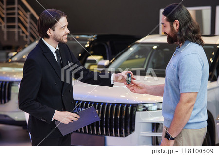 Side view of smiling male auto dealer in formal black suit handing smart key to adult man buying new vehicle at car dealership center 118100309