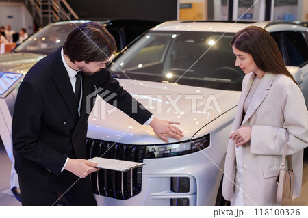 Side view of male auto dealer showing perfect polish of new luxury car to young female customer in elegant beige suit, while advising client on buying vehicle at car dealership center Side view of male auto dealer showing perfect polish of new luxury car to young female customer in elegant beige suit, while advising client on buying vehicle at car dealership center 118100326