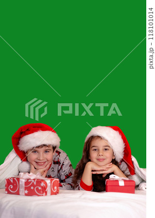 Two cute children, a girl and a boy, in pajamas and santa claus hats, on the bed 118101014