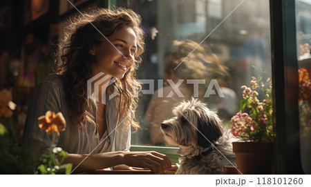 A smiling woman with curly hair sits with a dog in a sunny cafe by the window. 118101260
