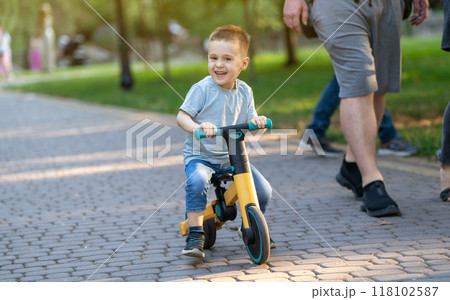 A cute smiling toddler boy of three or four years old rides a bicycle or balance bike in a city park on a sunny summer day. Toddlerhood and childhood concept. Selective focus A cute smiling toddler boy of three or four years old rides a bicycle or balance bike in a city park on a sunny summer day. Toddlerhood and childhood concept. Selective focus 118102587