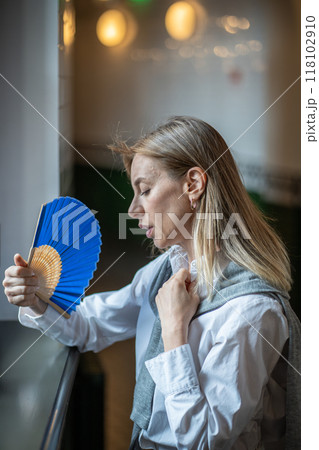 Overheated tired woman fans herself, cools down by window in office hall. Stuffy, no air conditioner 118102910