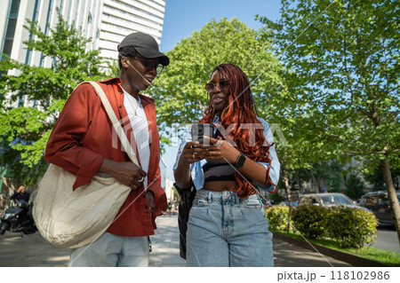 Stylish african american couple talking outdoor, stop walking on sidewalk looks at screen smartphone Stylish african american couple talking outdoor, stop walking on sidewalk looks at screen smartphone 118102986