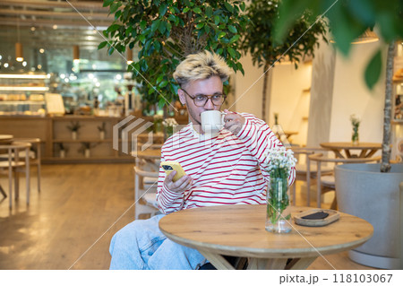 Reflective man sits with smartphone, drinks coffee and surfs internet, social media at cafe table 118103067