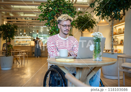 Focused disabled man in wheelchair works on laptop at cafe table. Freelancer working remotely online Focused disabled man in wheelchair works on laptop at cafe table. Freelancer working remotely online 118103086