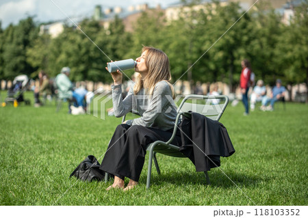 Relaxed woman drinking water from bottle relaxing in park on lawn sitting on folding chair. 118103352