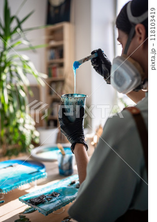 Rearview female artist assesses consistency, thickness of epoxy resin before starting artwork pour 118103419