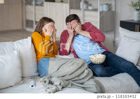 Scared family couple cover eyes with palms watching horrifying movie series sitting on couch at home 118103432
