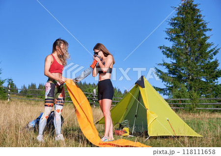 Two women hikers camping in mountains. Female tourists inflating the air mattress, wearing sportswear, hiking, making campsite. Concept of tourism and traveling. 118111658