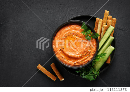 Htipiti - Greek red pepper and feta dip in black bowl with bread sticks and cucumbers, on dark background. top view, copy space 118111878