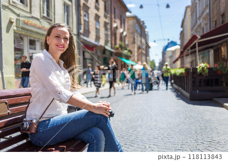 Young woman with the film camera sitting on a bench in a busy street, enjoying a sunny day Young woman with the film camera sitting on a bench in a busy street, enjoying a sunny day 118113843